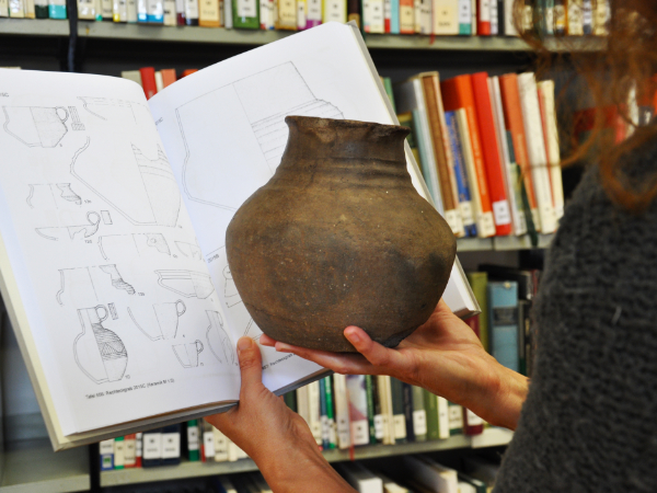 Person hält antike Vase in der Hand und vergleicht diese mit Aufzeichnungen in einem Buch.