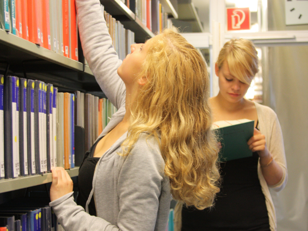 Zwei Studentinnen in der Bibliothek. Eine der Beiden nimmt ein Buch aus einem Regal, die andere liest