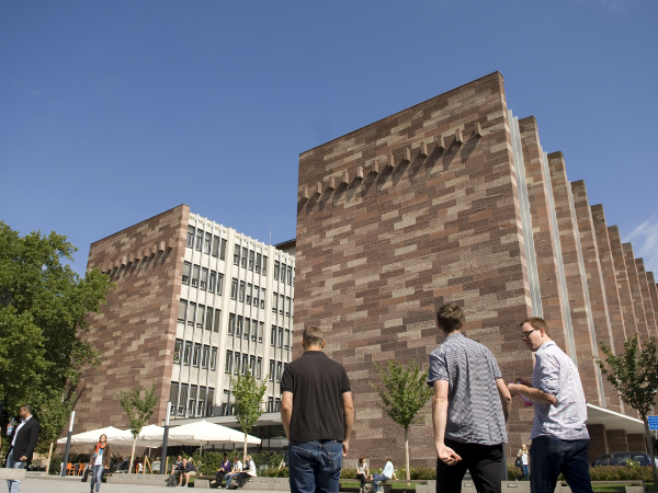 Im Vordergrund laufen drei Studenten über den Campus. Im Hintergrund ist das Kollegiengebäude II der Universität Freiburg zu sehen