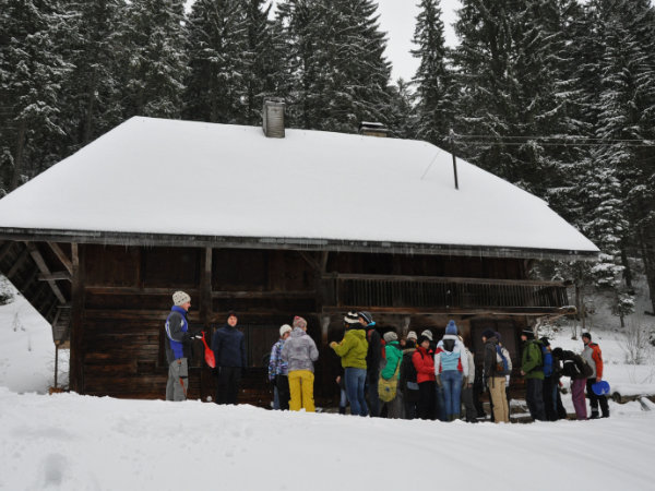 Schneebedeckte Hütte im Winter. Davor versammelt sich eine Gruppe Studierender