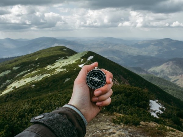 Eine Person hält einen Kompass aus Egoperspektive, im Hintergrund Berglandschaft